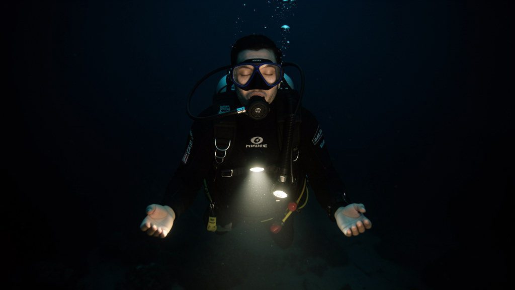 A scuba diver hovering motionless in dark water at night, dive light pointing downward, sense of flow state and deep peace